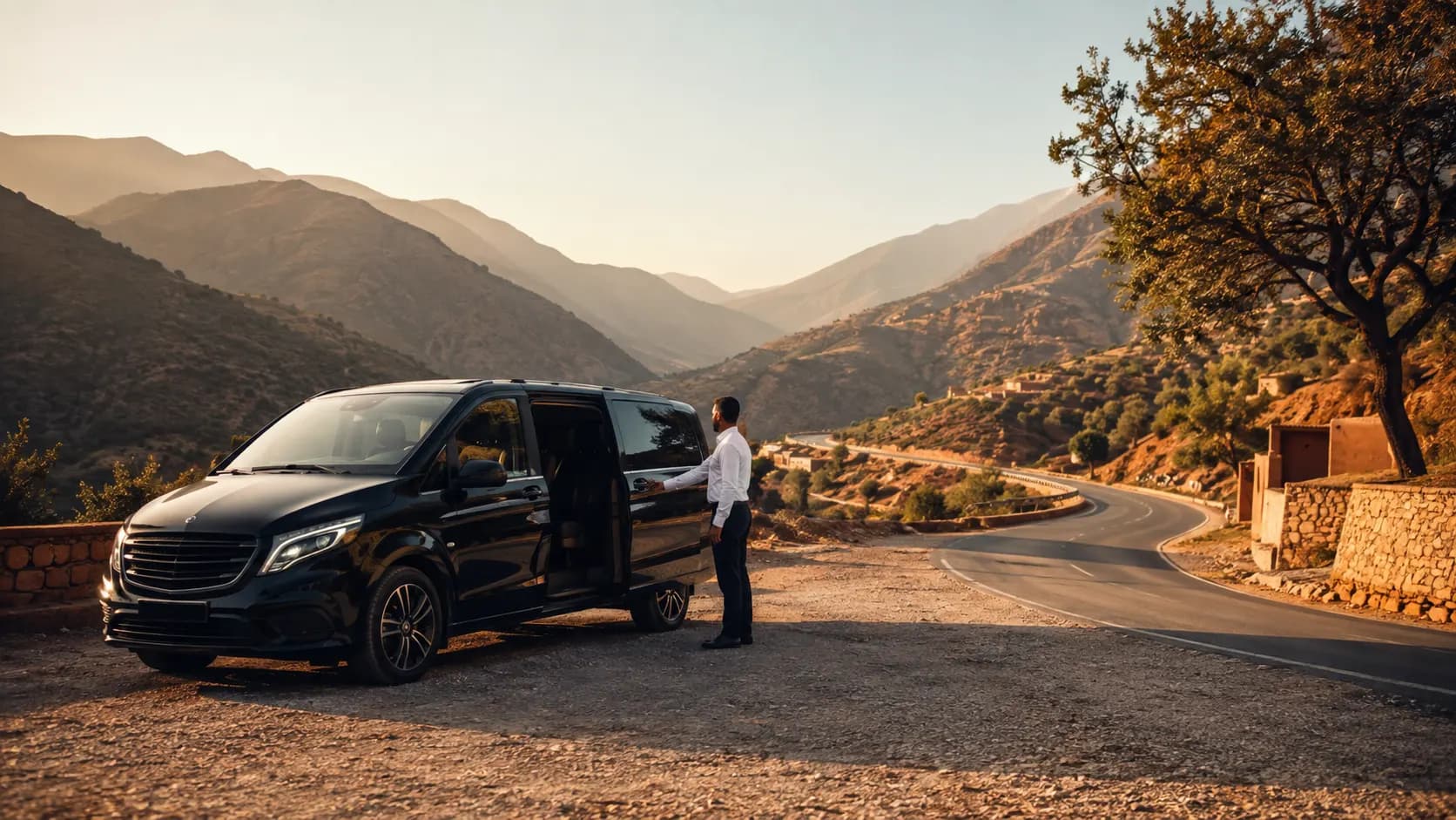 Professional private driver in Morocco opening the door of a black Mercedes Vito van on a scenic Atlas mountain road at sunset